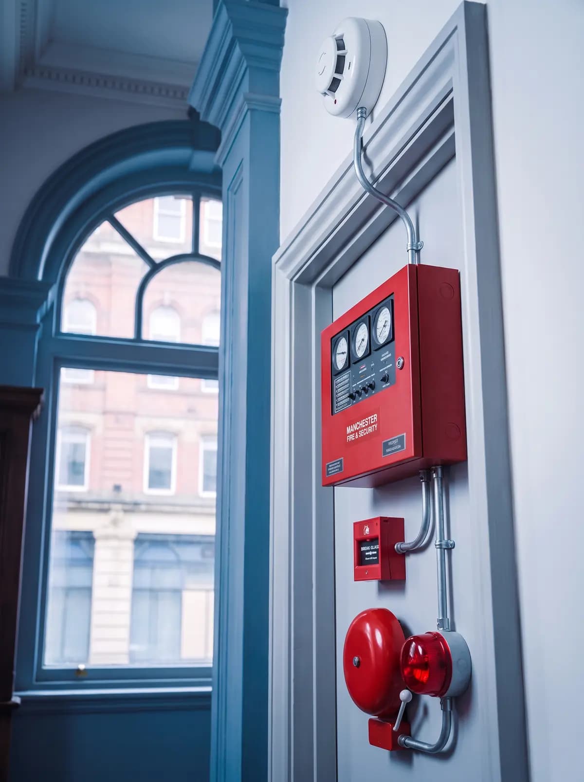 Red fire alarm panel, call point and bell on a wall beside a window