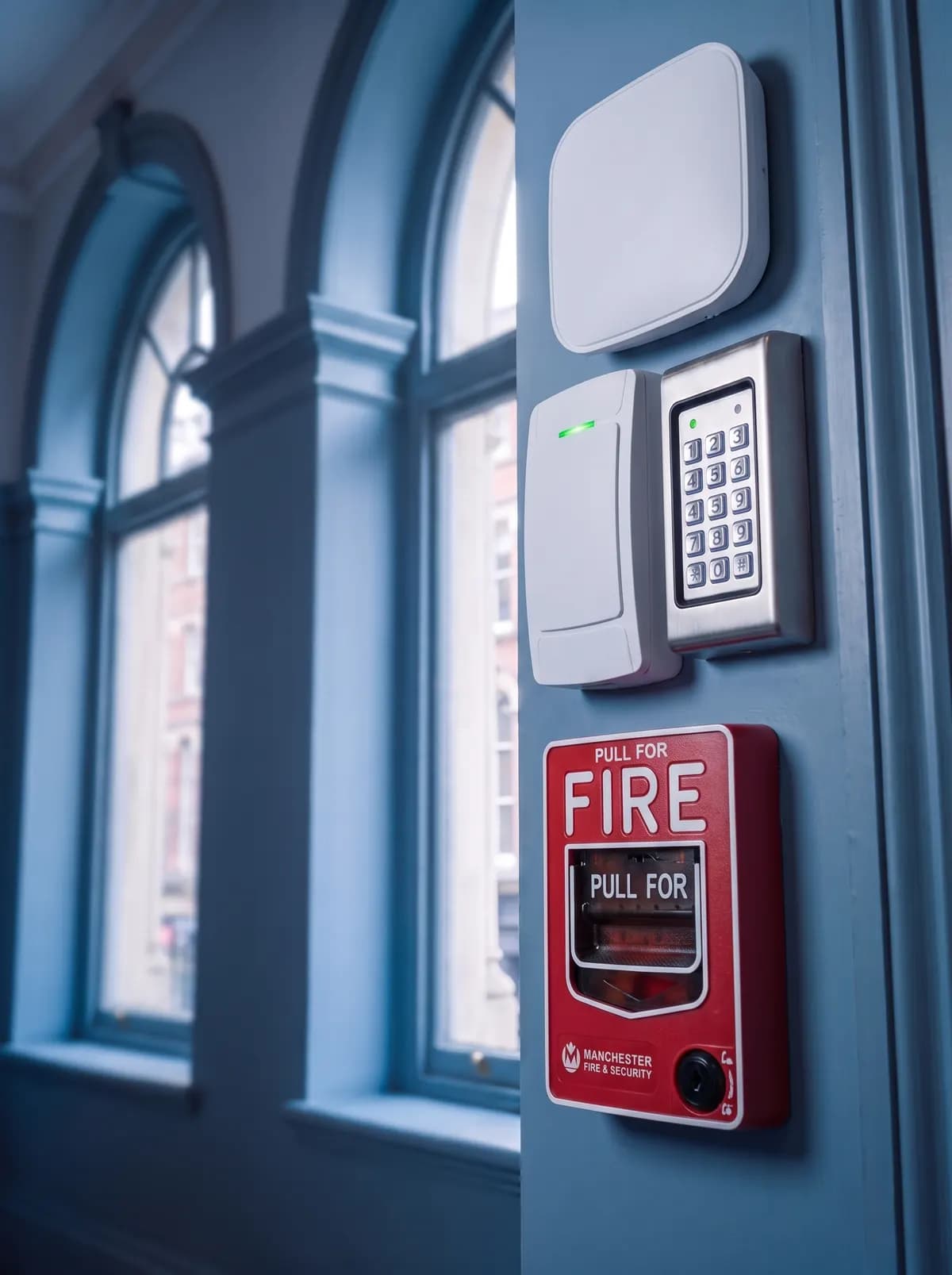 Red fire alarm pull station below a keypad and a reader on a wall